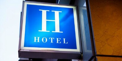 Outdoor illuminated sign with large letter H and “hotel” underneath, attached to a building.
