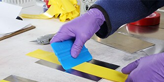 Gloved hands smooth out yellow tape on metal with 3M™ Hand Applicator PA1.
