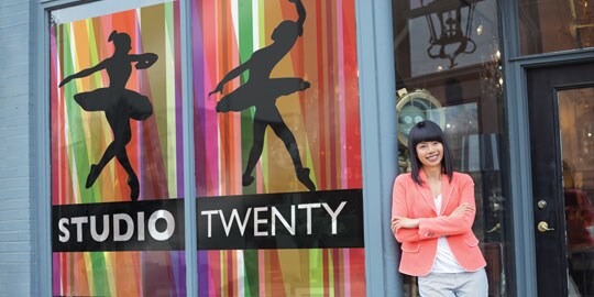 A woman leans against a storefront window wraps displaying dancers and the words studio twenty.
