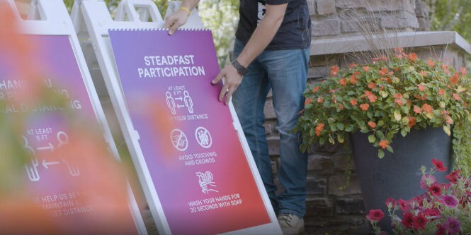 A person standing outdoors installing sign outlining COVID-19 guidelines on a sandwich-style board.
