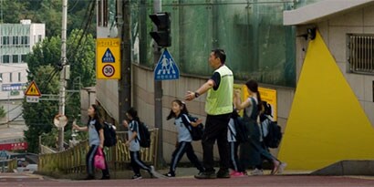 Children cross a street, directed by an adult in a fluorescent safety vest, in a city with mountains.
