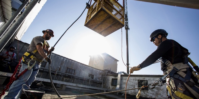 Image of two workers guiding hoses out of the way as a crane lowers a cage platform down to them
