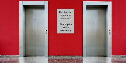 Two elevators with a sign in between reading “Only 1 person allowed in elevator! Wearing of a mask is mandatory.”
