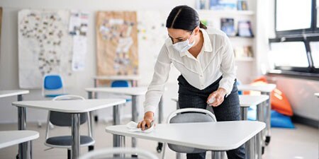 A woman with a mask on cleaning a school desk with 3M Instant Microbe Removing Wipes.

