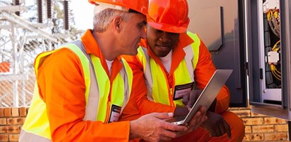 Two men wearing construction orange gaze at a laptop showing 3M training videos related to installing 3M electrical products.
