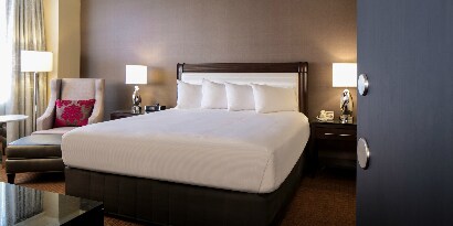 Hotel room with clean, tidy bed that has a white blanket, and lamps and armchair.
