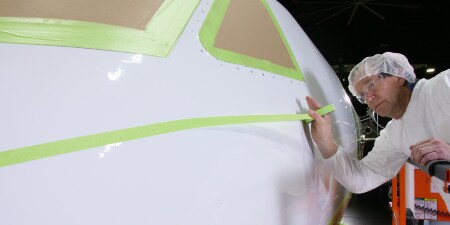 Worker applying masking tape to the nose of an aircraft whose cockpit windows are already masked

