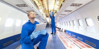 Image of an engineer with digital tablet looking up at wiring on ceiling of brightly lit, empty passenger jet.
