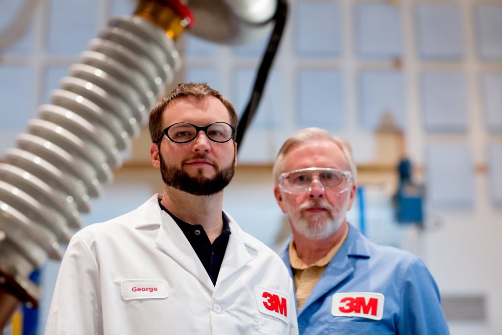 2 men in 3M lab coats standing in front of a transformer

