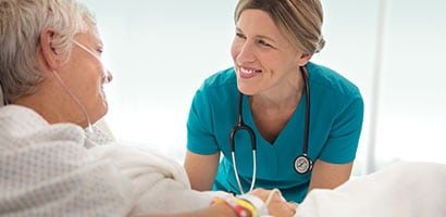 A female clinician wearing a stethoscope holding the hand of an elderly patient.
