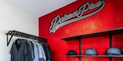 A display of shirts and caps is against a bright red wall with the Palmer Plus Apparel logo.

