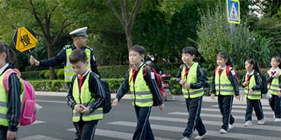 Schoolchildren wearing safety vests cross a crosswalk with an adult holding a school zone safety sign.
