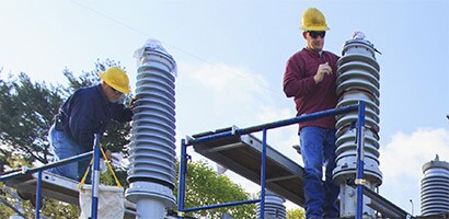 Two men in hardhats work high above the ground on high voltage joints and terminations.
